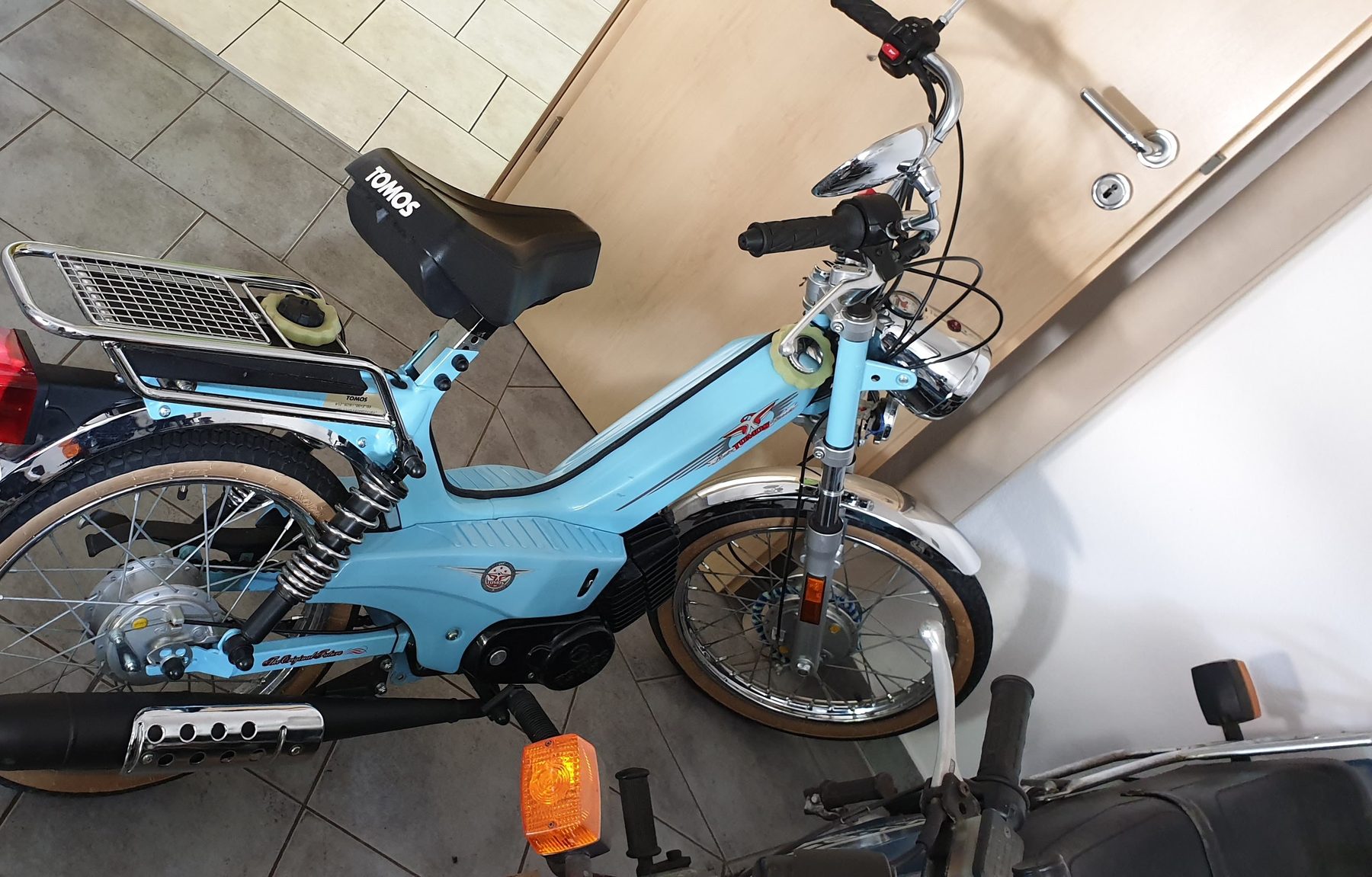 Hellblaues Tomos-Mofa mit Gepäckträger und Chromauspuff auf gefliestem Boden